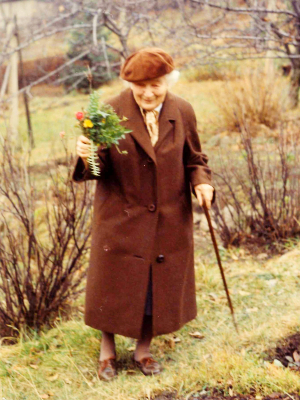 [Translate to Englisch (Intl):] Grete Wendt im braunen Mantel und mit Mütze mit einem bunten Blumenstrauß in der Hand. Sie steht in einem sonst herbstlichen Garten.