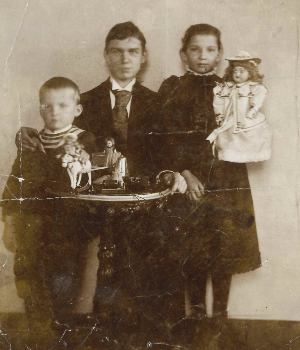 [Translate to Englisch (Intl):] Schwarz-Weiß-Aufnahme. Grete im dunklen Kleid und mit Puppe im Arm neben ihren Brüdern. Curt im Anzug, Johannes als kleiner Junge im Ringelpullover.